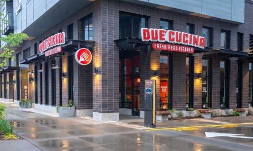 Exterior corner view of Due' Cucina Fresh Real Italian restaurant, featuring bold red signage on a dark brick building with warm interior lighting reflected on a wet sidewalk.