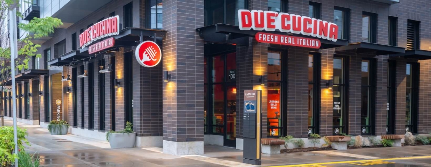 Exterior corner view of Due' Cucina Fresh Real Italian restaurant, featuring bold red signage on a dark brick building with warm interior lighting reflected on a wet sidewalk.
