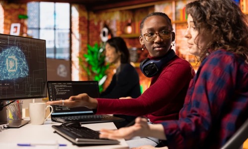 Two women collaborating at a desk in a modern office, one pointing at a computer screen displaying a digital AI brain interface while discussing the project.