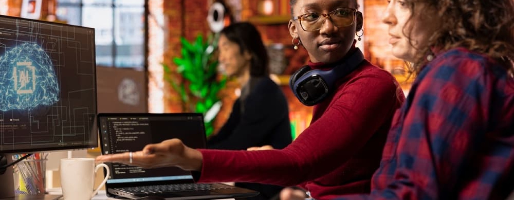 Two women collaborating at a desk in a modern office, one pointing at a computer screen displaying a digital AI brain interface while discussing the project.